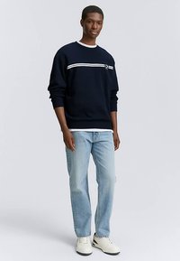 Marinblå sweatshirt med vita ränder över bröstet, kombinerad med ljusblå jeans och vita skor. Mjuk textur, rund halsringning.