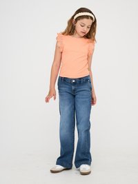 Pfirsichfarbener Ripp-Top mit gewellten Spitzenärmeln, kombiniert mit blauen, ausgestellten Jeans. Das Model trägt weiße Sneakers mit hellbraunen Akzenten.