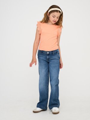 Breskva pletena majica s čipkastimi rokavi v obliki valov, v kombinaciji z modrimi širokimi denim hlačami. Model nosi bele teniske s svetlo rjavimi detajli.
