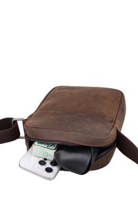 Sac bandoulière en cuir marron partiellement ouvert montrant un smartphone, un paquet de mouchoirs et un portefeuille noir à l'intérieur.