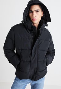 Giubbotto imbottito nero con chiusura a zip frontale, cappuccio e design trapuntato. Presenta polsini elastici e regolazione con cordoncino. Texture morbida e opaca.