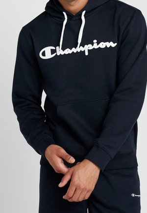 Navy hoodie featuring a white "Champion" logo, drawstring hood, front pocket, and ribbed cuffs and hem. Fabric appears soft and textured.