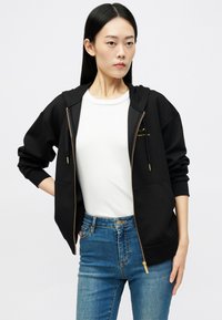 Jovem mulher vestindo um hoodie preto com fecho éclair por cima de uma camisa branca e jeans azuis, de pé contra um fundo branco simples.