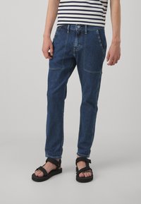 Los pantalones vaqueros de denim azul oscuro presentan un corte relajado, con costuras visibles, bolsillos laterales y un cierre de botón. Se llevan con sandalias negras.