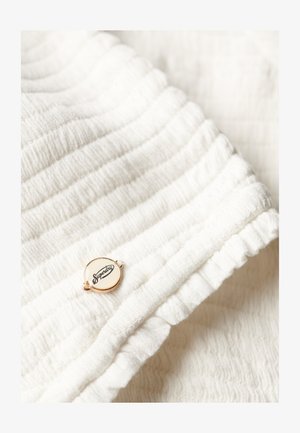 Witte gestructureerde stof met een verticaal ribbelpatroon, voorzien van een ronde gouden en crèmekleurige label met "Superdry"-merk.