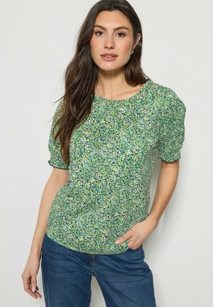 Frau mit langen braunen Haaren, die ein grünes, geblümtes Oberteil mit Puffärmeln und blaue Jeans trägt, steht mit einer Hand in der Tasche vor einem schlichten Hintergrund.