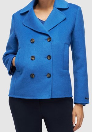 Femme portant un manteau en laine bleu vif à double boutonnage avec six boutons, poches latérales et grand col, assorti à un pantalon foncé.