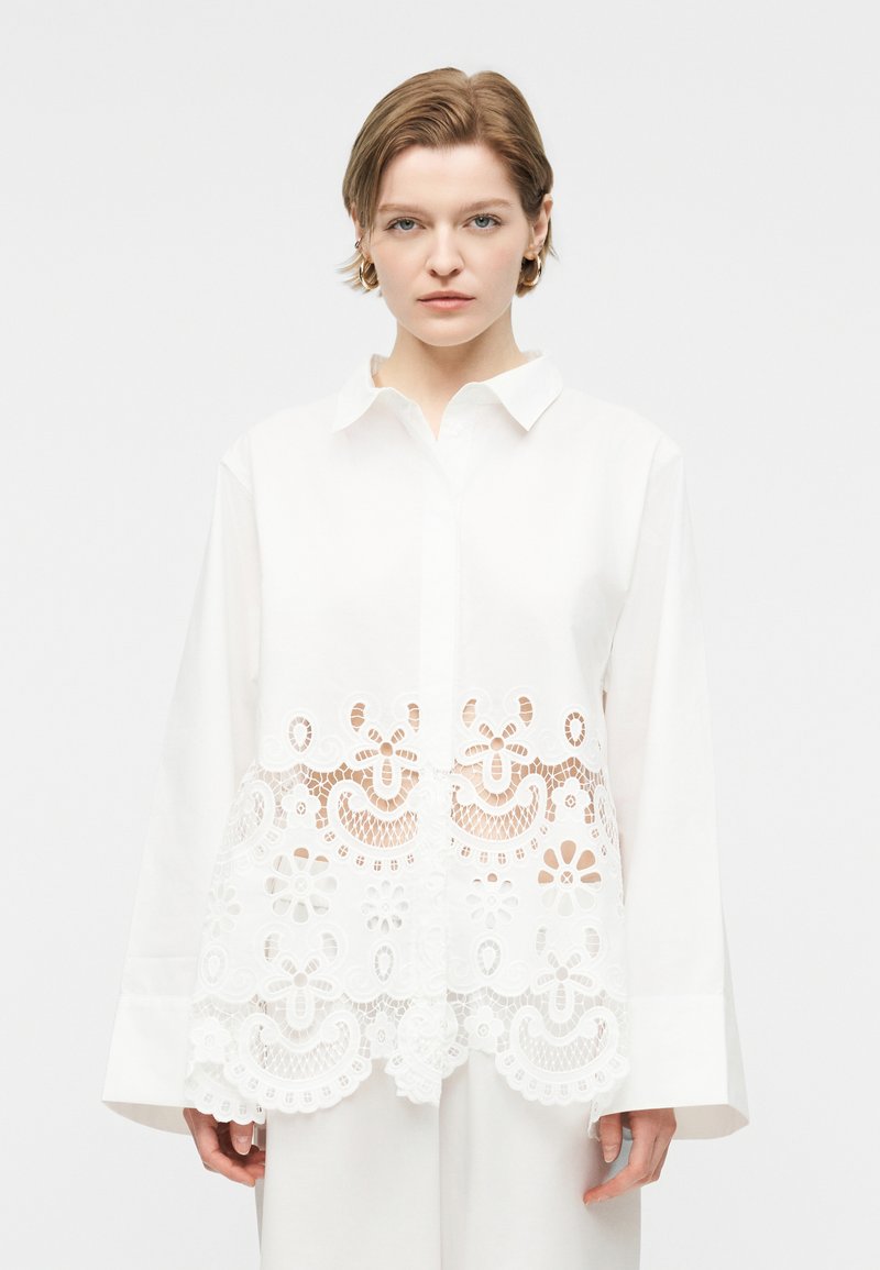 Γυναίκα που φοράει λευκό μπλουζάκι με μακριά μανίκια, γιακά και διακοσμητικά στοιχεία από δαντέλα με λουλούδια στο τελείωμα και τη μέση.