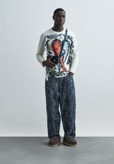 Μακρυμάνικη κρεμ μπλούζα με πολύχρωμο αφηρημένο σχέδιο, συνδυασμένη με φαρδιά σκούρα τζιν παντελόνια με υφασμένους μοτίβους και καφέ παπούτσια.