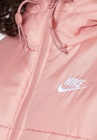 Gros plan sur une doudoune Nike rose avec fermeture éclair et cordons de capuche ajustables, mettant en avant le logo Nike brodé en blanc.