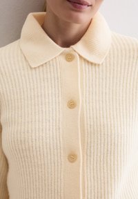 Beiger, gerippter Strickcardigan mit einem umgeschlagenen Kragen und fünf beigen Knöpfen auf der Vorderseite. Weiche Textur und lässiges Design.