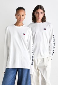 Witte longsleeves hebben een klein geborduurd logo en "TOMMY JEANS" gedrukt op de mouw. Modellen dragen casual jeans en broeken.
