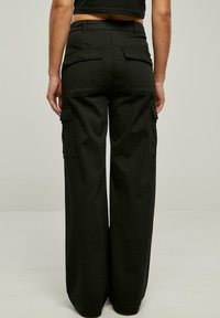 Nainen, joka on pukeutunut korkeavyötäröisiin mustiin leveälahkeisiin cargo-housuihin, joissa on läppäpussit, seisoo tasaisen vaalean taustan edessä.