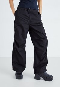 Pantalons cargo noirs à jambes larges avec une texture lisse, dotés de poches latérales et d'une coupe décontractée. Portés avec des sabots noirs à plateforme épaisse.