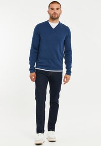 Marineblauer V-Ausschnitt-Pullover aus Rippstrick, kombiniert mit dunklen Jeans. Weißes T-Shirt unter dem Pullover, getragen mit hellen Sneakers.