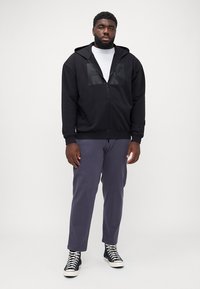 Zwarte zip-up hoodie met netpaneelaccenten, gedragen over een wit shirt. Gecombineerd met grijze broek en zwarte high-top sneakers.