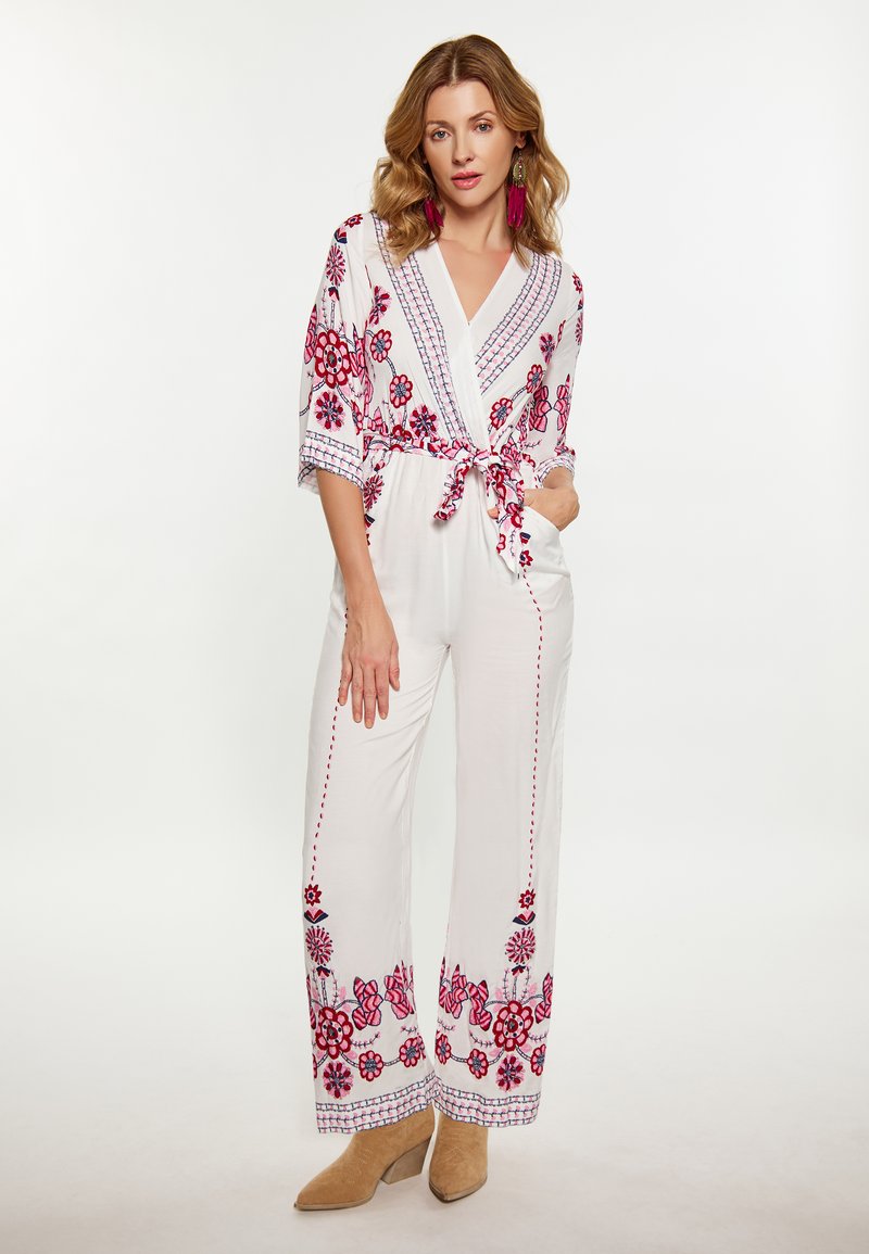 Salopetă albă cu broderie florală albastră și roșie, talie legată, mâneci 3/4 și pantaloni largi. Cizma cu toc maro completează ținuta.