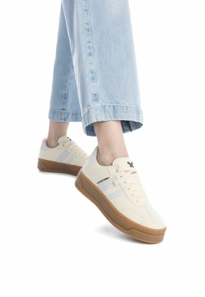 Zapatillas beige con una parte superior de tela texturada, franjas laterales blancas y suela de goma natural, mostradas con pantalones de mezclilla anchos de color azul.