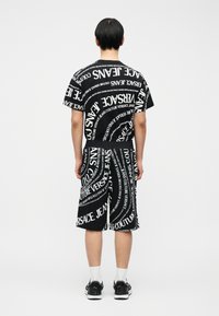 Άτομο που στέκεται με την πλάτη στην κάμερα, φοράει μαύρο σορτς και μαύρο t-shirt καλυμμένο με επαναλαμβανόμενο λευκό κείμενο Versace Jeans Couture.