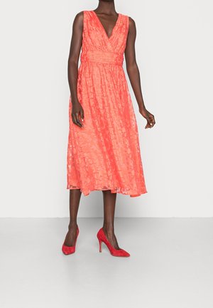 Femme portant une robe midi corail sans manches avec un décolleté en V et un motif texturé, associée à des chaussures à talons rouges.