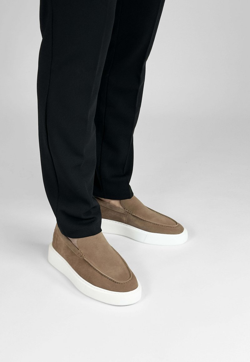 Bruine suède instap schoenen met een witte rubberen zool, voorzien van gestikte accenten en een gladde textuur, gecombineerd met zwarte broeken.