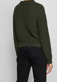 Mörkgrön stickad tröja med ett texturerat mönster, rund halsringning och sänkta axlar. Kombinerad med svarta byxor som visar en avslappnad passform.