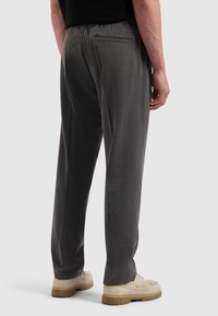 Man in donkere grijze broek en beige schoenen, gezien van achteren tegen een effen lichte achtergrond.
