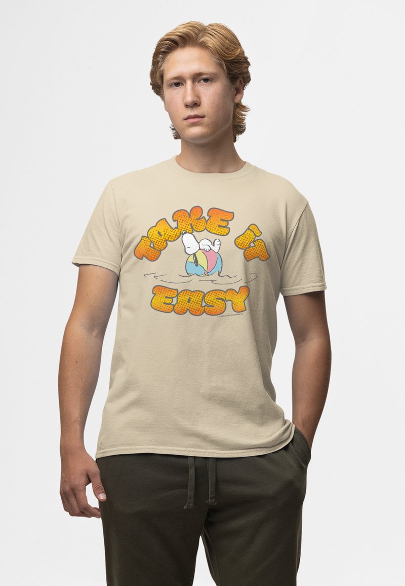 Jeune homme aux cheveux blonds portant un t-shirt beige avec Snoopy flottant sur un anneau de piscine et le texte "Take it easy" en lettres bulles orange.