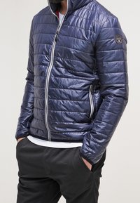 Giacca puffer blu scuro con design trapuntato, zip completa, colletto alto e patch del logo sulla manica, abbinata a pantaloni neri e una camicia bianca.