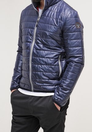 Marinblå puffjacka med quiltad design, full dragkedja, hög krage och logopatch på ärmen, matchad med svarta byxor och en vit skjorta.