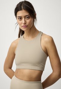 Beige sportsbh med høj hals, sømløst design og blødt, strækbart stof. Har en minimalistisk silhuet og let tekstur.