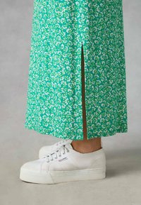 Grön blommig kjol med en slits på sidan, tillverkad av lättviktsmaterial, tillsammans med vita kanvas sneakers med en texturerad sula.