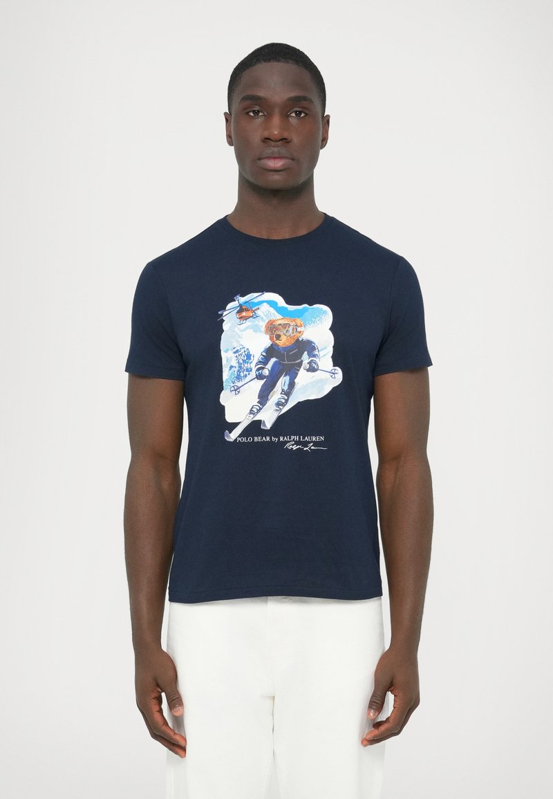 Majica tamnoplave boje od pamuka s grafikom medvjeda koji skija na bijeloj pozadini te tekstom na dnu. Kratki rukavi, okrugli izrez.