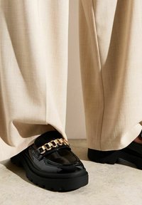 Mocassini neri in vernice con suole chunky, caratterizzati da un accento in catena dorata sulla parte anteriore, abbinati a pantaloni a gamba larga beige chiaro.