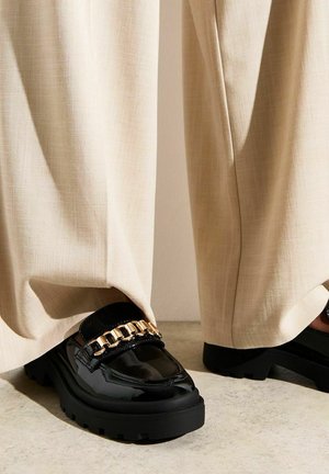 Mocasines de charol negros con suelas gruesas, adornados con una cadena dorada en la parte frontal, combinados con pantalones palazzo de un beige claro.
