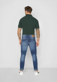 Világoskék mosású farmernadrág, tapered fit szabással, két hátsó zsebbel és bézs bőrbélyegzővel. Sötétzöld polo pólóval párosítva.