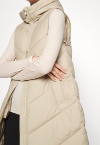 Beige gepolsterte Weste mit hohem Kragen, diagonalen Nähten, einem Reißverschluss vorne und ohne Ärmel. Weiches, leichtes Material.