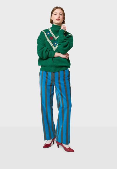 Zeleni pleteni pulover s visokim ovratnikom i cvetnim vzorcem, v kombinaciji z modrimi prugastimi hlačami in rdečimi visokimi petami.
