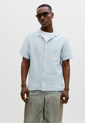 Mężczyzna o krótkich włosach i okularach przeciwsłonecznych, ubrany w jasnoniebieską, wzorzystą koszulę z krótkim rękawem na białej koszulce oraz luźne szare spodnie.