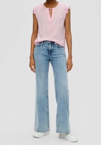 Femme portant un chemisier sans manches rose avec des bords en dentelle, un jean large bleu clair et des baskets blanches, debout devant un fond uni.