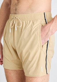 Shorts de bain beiges en tissu léger, dotés d'une taille élastique, d'un cordon de serrage, d'une pochette latérale et de rayures noires et blanches sur les côtés.