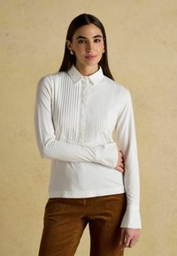 White long-sleeve shirt featuring a textured, pleated front panel and a collared neckline, paired with brown corduroy pants.