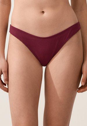 Ženska v bordo rjavih bikini spodnjicah z teksturo, prikazana od sredine trupa do zgornjega dela stegen, stoji pred nevtralnim ozadjem.