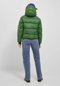 Grön puffjacka med huva, med sömmade paneler. Buren över blå denimjeans, med en avslappnad keps på huvudet.