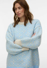 Femme aux cheveux bruns portant un pull en tricot à motifs bleu clair et crème à manches longues, debout les bras croisés, regardant vers la gauche.