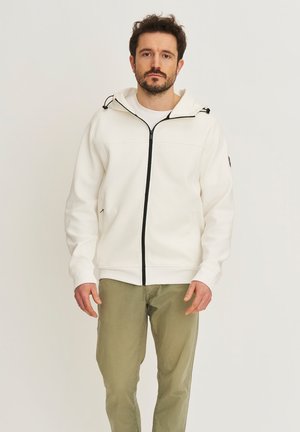 Man met donker haar, gekleed in een wit vest met rits en olijfgroene broek, staand tegen een effen lichte achtergrond.