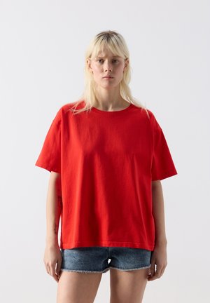 Blonde woman wearing a loose bright red short-sleeve t-shirt and denim shorts standing against a plain light background.