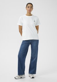Camiseta de algodón blanca de manga corta y cuello redondo, con un pequeño emblema de corazón negro en el pecho, combinada con unos vaqueros anchos de color azul.