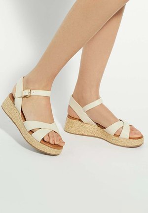 White faux leather wedge sandals with a woven jute platform, cross-straps, and ankle buckle. Smooth texture and open-toe design.