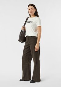 Biała koszulka z nadrukiem "STUDIO", oliwkowe spodnie o szerokich nogawkach oraz czarne buty. Modelka trzyma brązową skórzaną torbę typu tote. Prosty design.